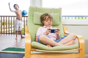 Two children, one using a tablet and one with a ball. A concept for too much screen time for children.